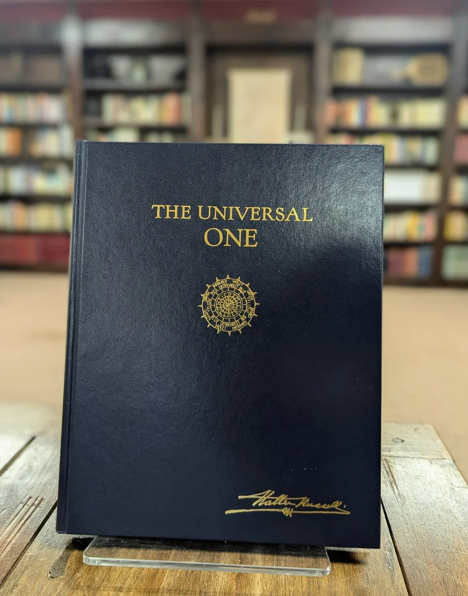 The Universal One — a navy hardcover with gold lettering, the Cosmic Clock seal, and Walter Russell's signature — standing on a wooden table in front of bookshelves.