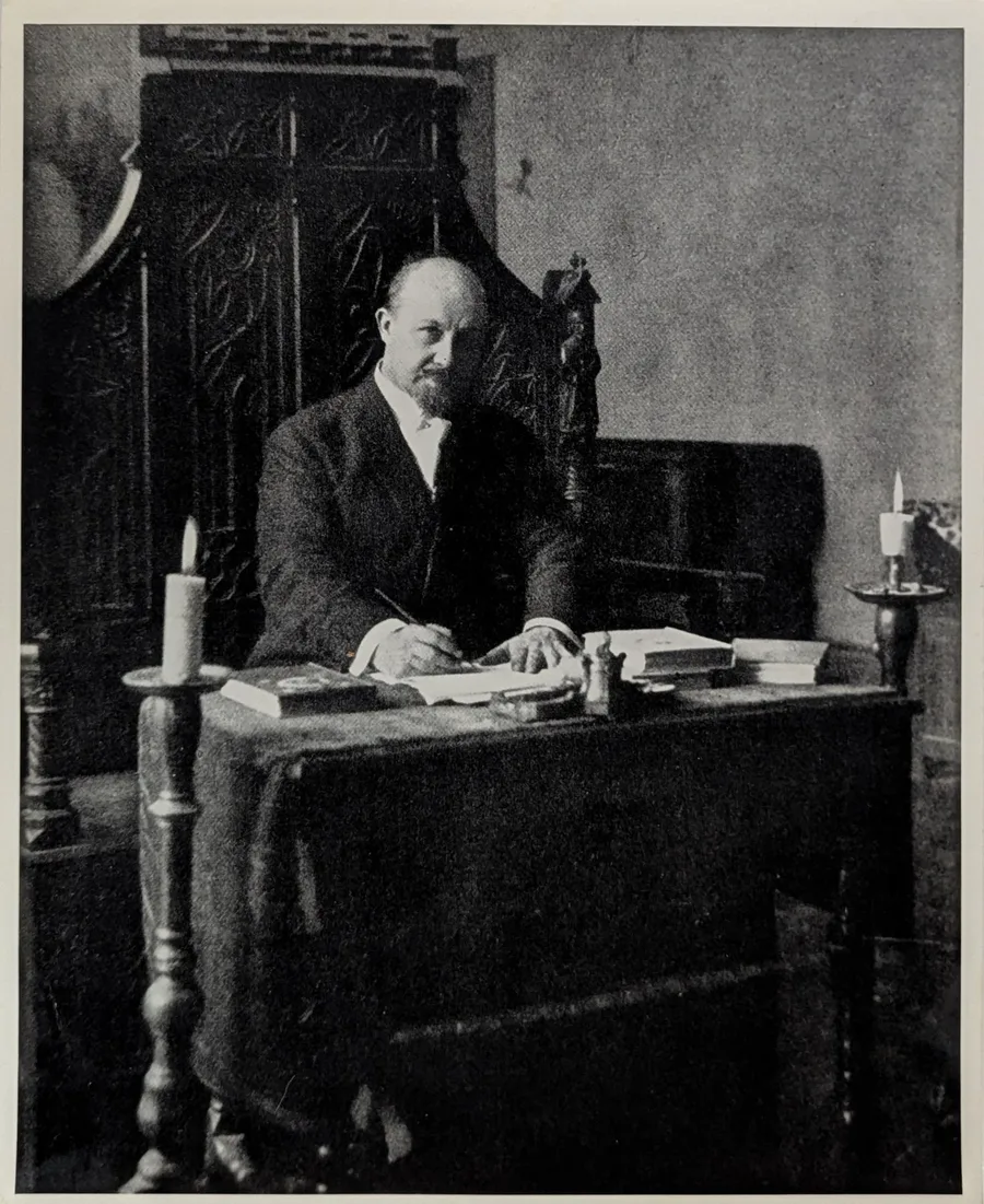 Walter Russell writing at a desk by candlelight, seated in a carved high-backed chair with books and papers around him.